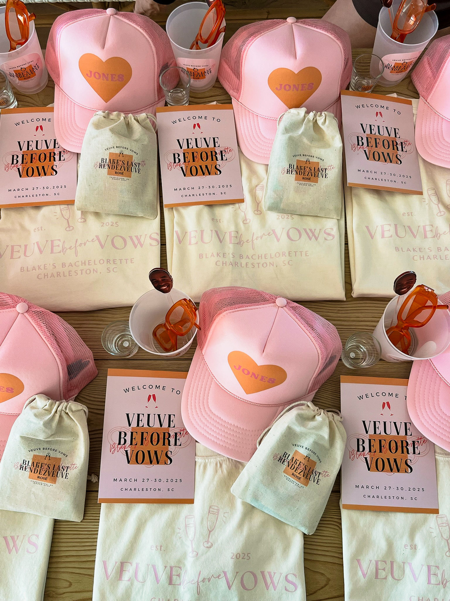 Flat-lay of pink bachelorette swag in Charleston: pink mesh trucker caps with heart decals, themed welcome cards, drawstring favor bags, orange sunglasses and shot glasses.