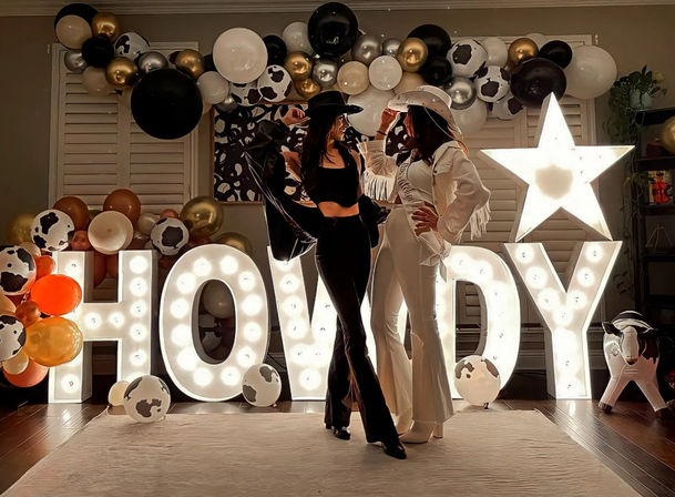Two people in black and white cowgirl outfits and hats pose in front of a giant illuminated HOWDY marquee and star, surrounded by black, white, gold and cow-print balloon garland for a western-themed indoor party backdrop.