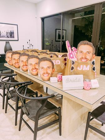 Bachelorette party gift setup on a modern dining table: rows of personalized burlap tote bags with oversized smiling male face cutouts, pink monogram tumblers, ‘Bride’ clutch, sunglasses and a foam finger.