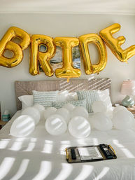 Sunlit bedroom with gold foil 'BRIDE' balloons above a bed scattered with white balloons, patterned pillows, lamp and a tray — wedding morning setup.
