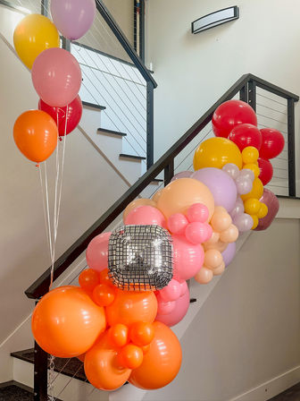 Vibrant balloon garland in orange, pink, yellow, red and lavender with a silver disco foil balloon draped along a modern indoor staircase railing — festive party decor