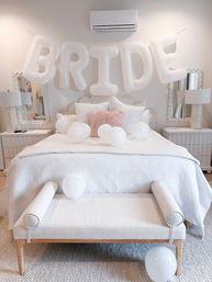 Bright neutral bridal suite bedroom with oversized white 'BRIDE' balloons above a quilted white bed, scattered white balloons, a blush pink accent pillow, mirrored nightstands and a light-wood bench