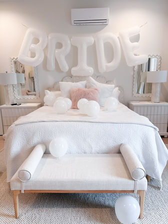 Bright neutral bridal suite bedroom with oversized white 'BRIDE' balloons above a quilted white bed, scattered white balloons, a blush pink accent pillow, mirrored nightstands and a light-wood bench