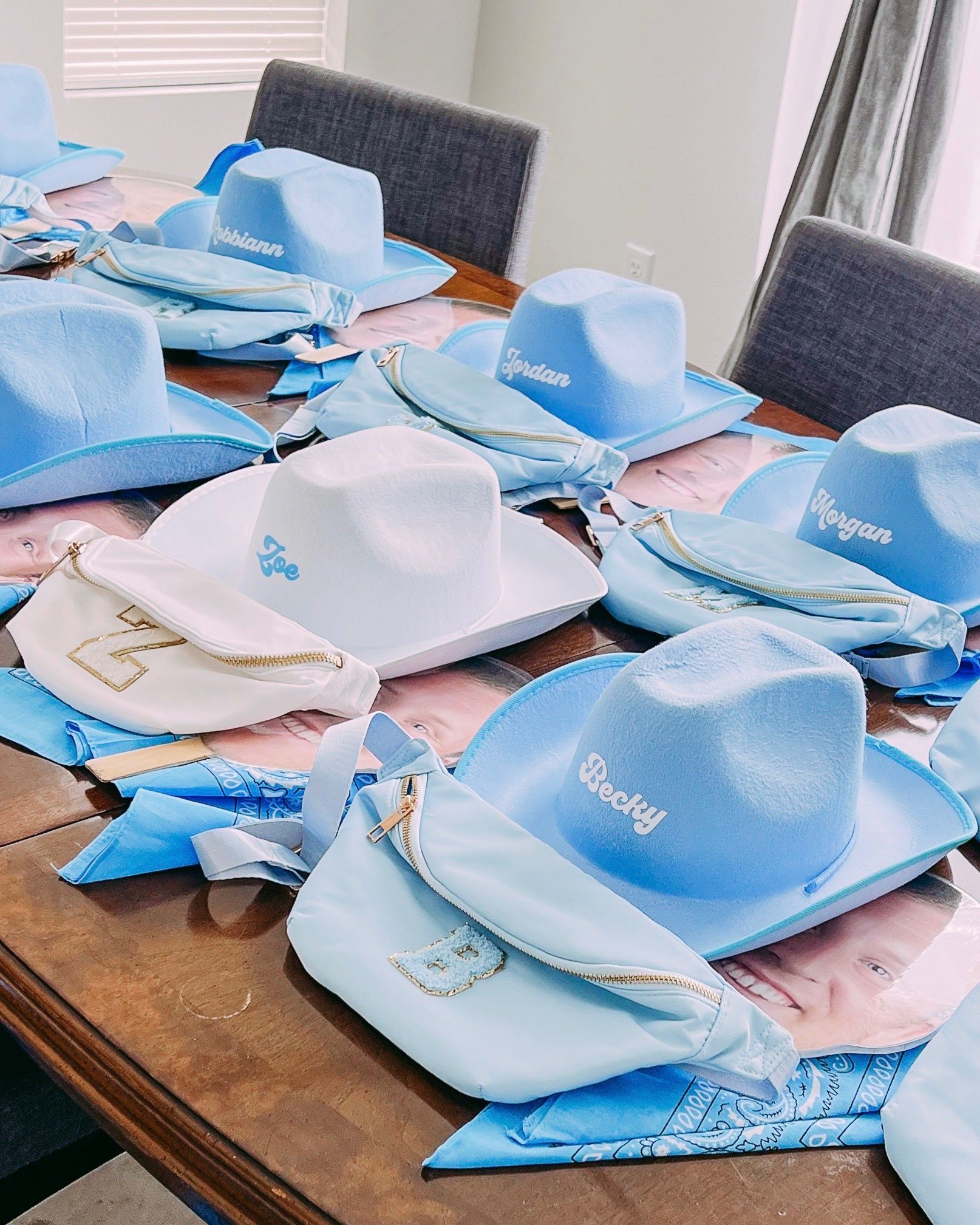 Pastel blue and white personalized cowboy hats with matching fanny packs and bandanas neatly arranged on a wooden dining table as western-themed party favors, ready for guests.