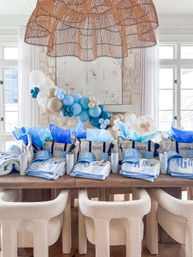 Sunlit coastal party setup with blue-and-white favor bags, personalized towels and 30A caps, tissue paper and tumblers on a wooden table, cream boucle chairs, a blue-and-white balloon garland and woven rattan pendant light.