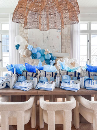 Sunlit coastal party setup with blue-and-white favor bags, personalized towels and 30A caps, tissue paper and tumblers on a wooden table, cream boucle chairs, a blue-and-white balloon garland and woven rattan pendant light.