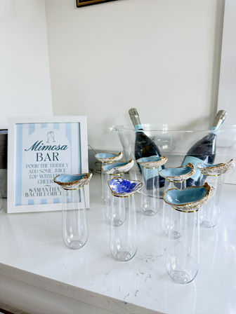 Coastal-inspired mimosa bar on a white marble countertop: framed 'Mimosa Bar' sign, two chilled sparkling wine bottles in a clear ice bucket, and tall flutes topped with blue-and-gold oyster-shell rims.