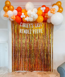 Indoor celebration photo backdrop with shimmering gold fringe curtain and a festive orange, white, and gold balloon garland with small balloon flower clusters.