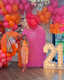 Vibrant indoor 21st birthday scene with a person in an orange sequin dress posing in front of a pink backdrop, surrounded by pink and orange balloon clusters, floral accents, a champagne-shaped balloon and illuminated marquee '21' lights.