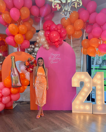 Woman in orange sequin midi dress posing indoors at a vibrant pink-and-orange balloon arch photo backdrop with foil champagne balloon, floral cluster and illuminated '21' marquee for a 21st birthday