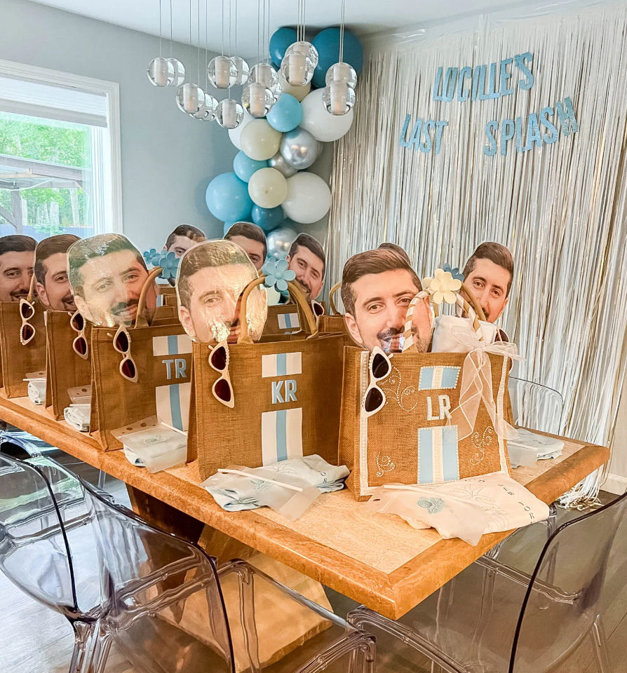Indoor celebration table with blue-and-white balloon garland and metallic fringe backdrop, row of burlap gift bags topped with oversized face cutouts and sunglasses, clear acrylic chairs and folded napkins — playful party decor.
