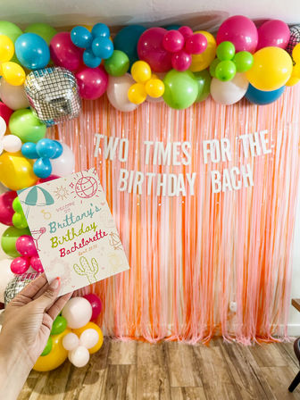 Indoor birthday bachelorette party scene with pink fringe backdrop, white letter banner, colorful balloon garland and a hand holding a pastel party invitation over wood flooring.