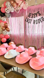 Pink rodeo-themed bachelorette setup: metallic pink fringe backdrop with a 'Last Rodeo' banner, pink/rose-gold/cow-print balloon garland, and a row of pink cowboy hats with a white "Bride" hat on the table.