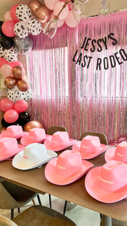 Pink rodeo-themed bachelorette setup: metallic pink fringe backdrop with a 'Last Rodeo' banner, pink/rose-gold/cow-print balloon garland, and a row of pink cowboy hats with a white "Bride" hat on the table.