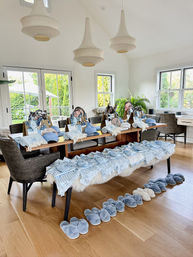 Sunlit modern dining room with wooden table arranged for a party: wicker gift bags, coordinated blue-striped robes and caps on chairs, a bench of matching outfits, and a neat row of fluffy blue slippers on the hardwood floor beneath pendant lights and large windows overlooking a green garden.
