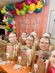 Indoor birthday party setup in a living room with a colorful balloon garland and pink fringe backdrop, table of burlap gift bags topped with oversized face cutouts, sunglasses, wine glasses and party favors.