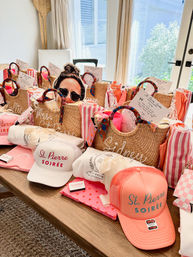 Beachy straw tote gift bags arranged on a dining table, each filled with pink-striped towels, scarves on the round handles, note cards and small treats, surrounded by coral and white trucker hats and matching pouches — colorful girls’ soiree welcome gifts.