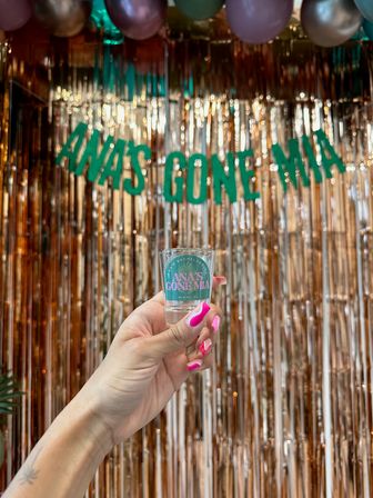Hand with hot-pink manicure holding a shot glass against a teal-letter name banner and metallic rose-gold fringe backdrop, balloons overhead — Miami party vibe