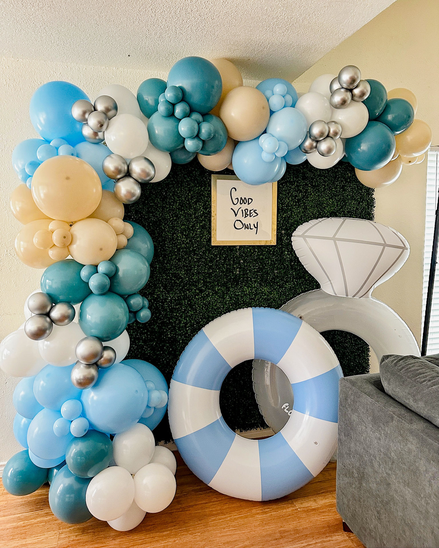 Playful indoor party backdrop: blue, teal, white, gold and silver balloon garland framing a boxwood photo wall with a 'Good Vibes Only' sign and oversized inflatable lifebuoy and diamond ring props.