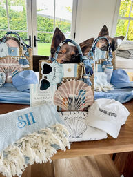 Hamptons-style beach gift bags and coastal accessories on a table — straw totes, monogram towel, caps, fans, and a small brown dog peeking from a tote