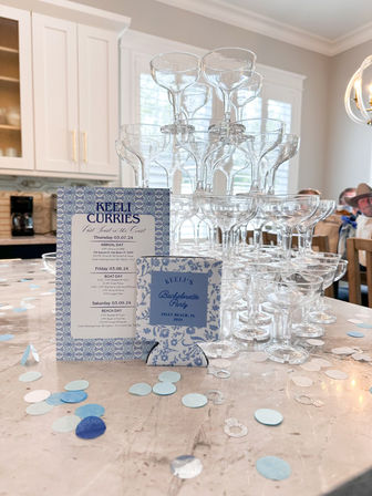 Blue-and-white bachelorette party invites and itinerary on a marble kitchen island with a stacked champagne coupe tower, scattered blue confetti and silver ring decorations in a bright coastal Inlet Beach, FL kitchen