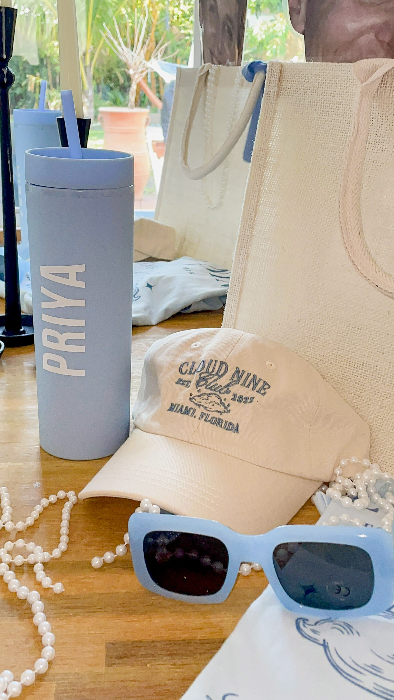 Pastel blue travel tumbler, cream baseball cap, light-blue square sunglasses, pearl necklace and woven tote arranged on a wooden table by a sunlit window — beach-ready Florida summer accessories.