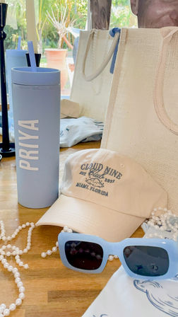 Pastel blue travel tumbler, cream baseball cap, light-blue square sunglasses, pearl necklace and woven tote arranged on a wooden table by a sunlit window — beach-ready Florida summer accessories.