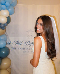 Smiling bride-to-be in a beaded white wedding dress and veil poses beside a blue-and-cream balloon arch in front of an event backdrop reading "The Hamptons".