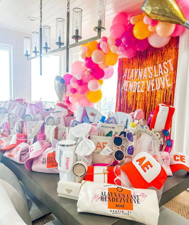Vibrant bachelorette favor spread in Destin, FL — pink and orange balloon arch, shimmering fringe backdrop, totes, sunglasses, caps and rosé-themed goodies arranged on a sunlit table.