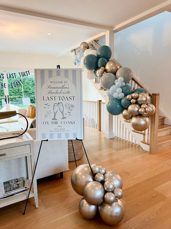 Coastal-themed bachelorette party welcome sign on an easel in a bright modern home, with teal and metallic gold balloon garlands cascading down a staircase.