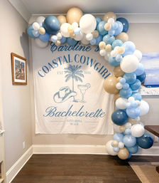 Santa Rosa Beach-themed bachelorette backdrop with palm tree, cowboy hat and cocktail illustration framed by a blue, white and tan balloon garland arch in a cozy indoor space