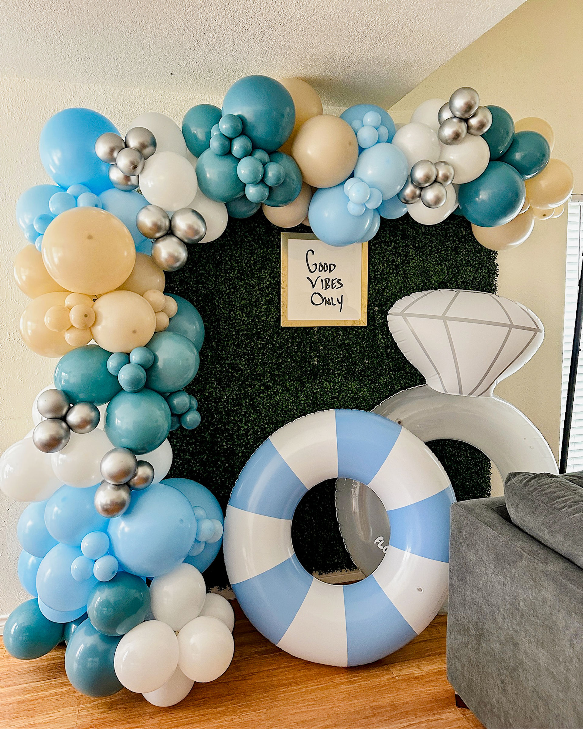 Indoor party photo backdrop: teal, light-blue, beige, white and silver balloon arch framing a boxwood hedge panel with framed "Good Vibes Only" sign, paired with a blue-and-white inflatable pool ring and oversized diamond ring prop on hardwood next to a gray sofa.