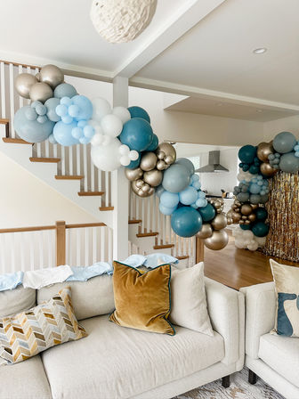 Playful blue, white, and metallic gold balloon garland draped along a staircase and archway in a modern open-concept living room with a light sofa and velvet throw pillows.