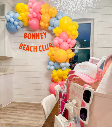 Cheerful pastel balloon arch (pink, yellow, blue) framing an indoor coastal-themed party setup in a modern farmhouse kitchen with an orange letter banner and table of party favors.