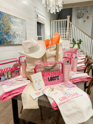 Bachelorette party welcome gifts on a coastal home's dining table: white 'BRIDE' straw hat on a canvas tote, pink clear pouches labeled bikinis, personalized tumblers, and matching pink-and-white tees and towels.