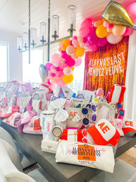 Bachelorette party gift table in Destin, FL with pink, orange and gold balloon garland, metallic fringe backdrop and themed favor bags, caps and sunglasses.