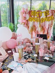 Fun indoor bachelorette party setup in a glass‑walled room with garden view: pastel pink, white and gold balloon garland and metallic fringe backdrop with gold letter balloons spelling a bachelorette bash; table strewn with glitter tumblers, pink cups, heart-shaped sunglasses, cozy slippers, confetti and tulle favors.