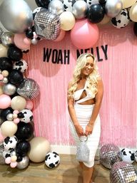 Smiling woman in a white cutout dress holding a sparkly silver clutch at a pink fringe photo backdrop reading “WOAH NELLY,” framed by a pink, black, silver and cow-print balloon arch with disco-ball props.