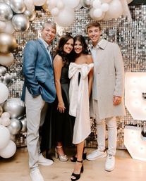 Four adults smiling and posing at an indoor celebration photo wall with a silver sequin backdrop, white and metallic balloon arch, lit marquee number, and a woman in the center wearing a white bow-front dress.