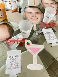Miami bachelorette party props on a glass table: oversized face cutouts, white plastic cups, pink martini cutout, heart-shaped red sunglasses, clear martini glass and printed can coolers.