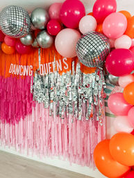 Bright disco-themed photo backdrop with pink and orange fringe, silver tassels, mirrored disco balls and a balloon arch around a 'DANCING QUEENS' banner