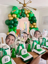 Home dining room party setup with green and gold balloon arch, a wooden table lined with white tote favor bags, green trucker hats, bandanas, mini megaphones and oversized smiling face cutouts for a festive celebration.