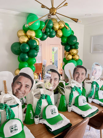 Home dining-room party setup with green and gold balloon arch over the doorway; wooden table lined with camp-themed favor bags, green-and-white trucker hats, bandanas, mini megaphones, tumblers and oversized smiling face cutouts.