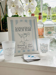 Beach-themed bachelorette bar display in the Hamptons: framed pastel-striped sign, white orchids, reusable cups, coasters and liquor bottles on a sunlit countertop.