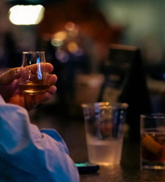 Close-up of a hand holding a whiskey glass on a dimly lit bar counter with a plastic cup and lemon cocktail nearby, warm bokeh lights creating a cozy nightlife vibe.