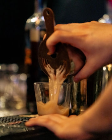 Close-up of hands pouring a creamy cocktail through a strainer into a short glass on a dimly lit bar, warm bokeh lights in the background.