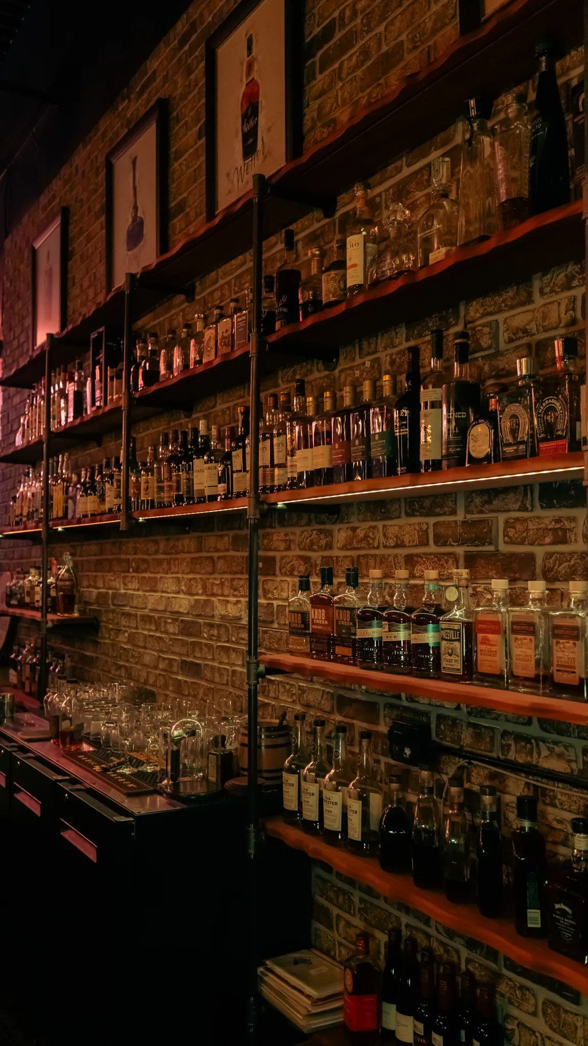 Cozy urban bar interior — warm-lit brick wall with wooden shelves and industrial pipes lined with whiskey and liquor bottles, glassware and bar tools.