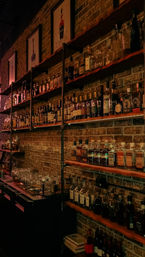 Cozy urban bar interior — warm-lit brick wall with wooden shelves and industrial pipes lined with whiskey and liquor bottles, glassware and bar tools.