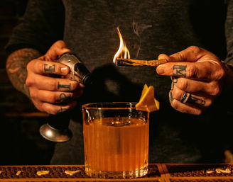 Tattooed hands of a bartender torching a cinnamon stick over an Old-Fashioned–style cocktail with an orange peel on a dim bar counter