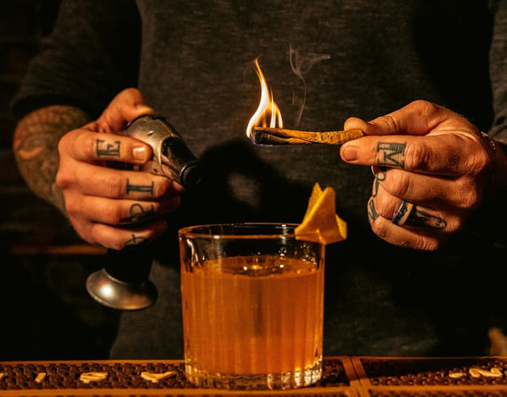 Tattooed hands of a bartender torching a cinnamon stick over an Old-Fashioned–style cocktail with an orange peel on a dim bar counter